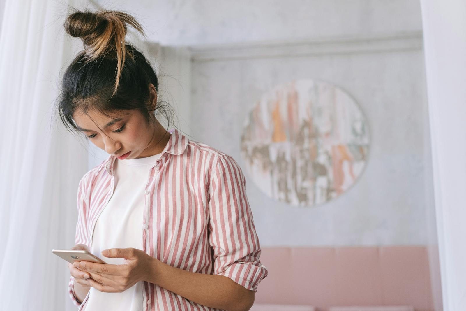 A young woman in a striped shirt using her smartphone indoors with soft decor and a relaxed atmosphere.