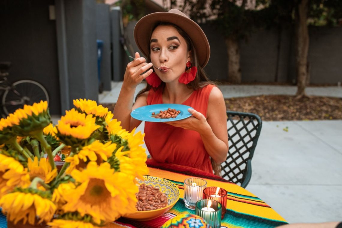 A Hispanic woman savoring a plate of beans in a vibrant outdoor setting with sunflowers.