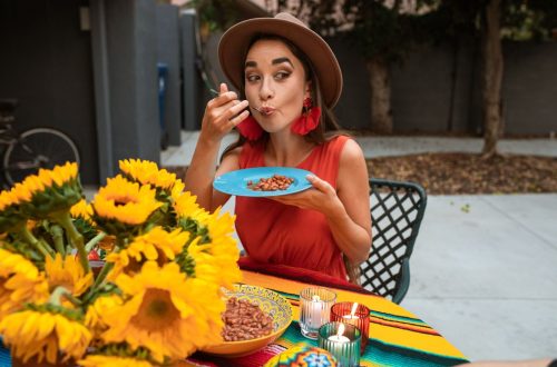 A Hispanic woman savoring a plate of beans in a vibrant outdoor setting with sunflowers.